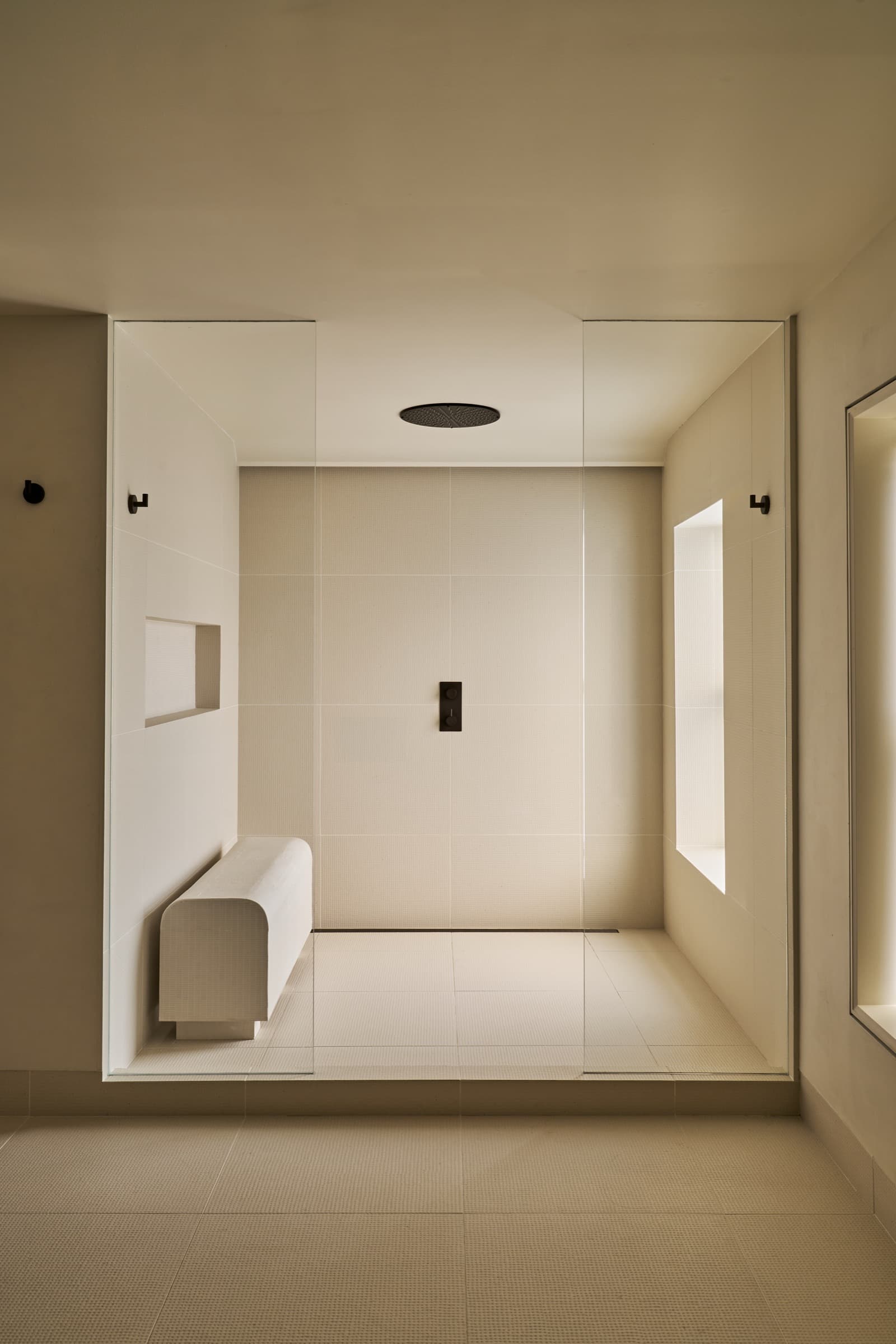Minimalist walk-in shower with pale tiled surfaces, built-in bench, and dark fixtures