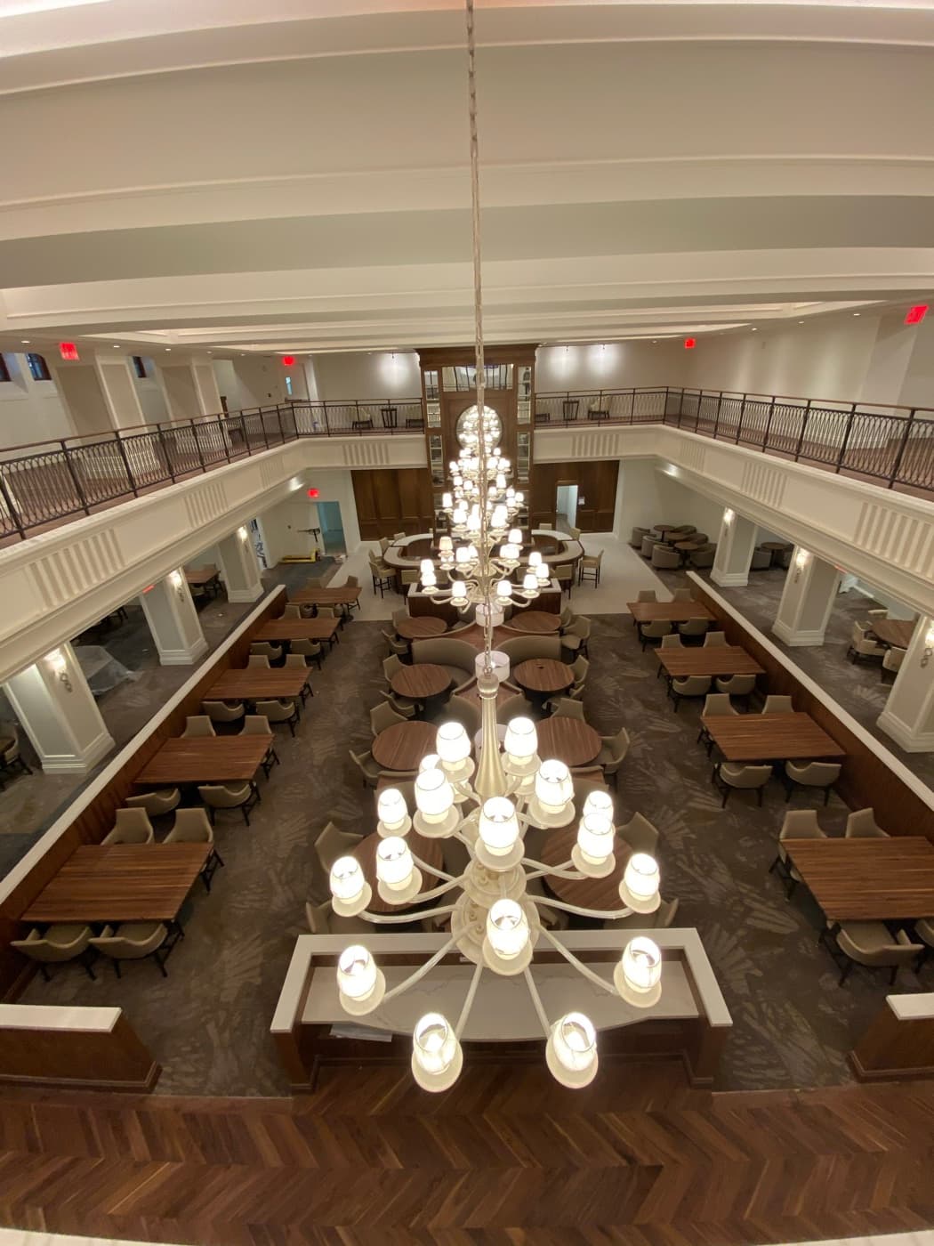 Double-height dining hall featuring a grand tiered chandelier, mezzanine railings, and timber tables