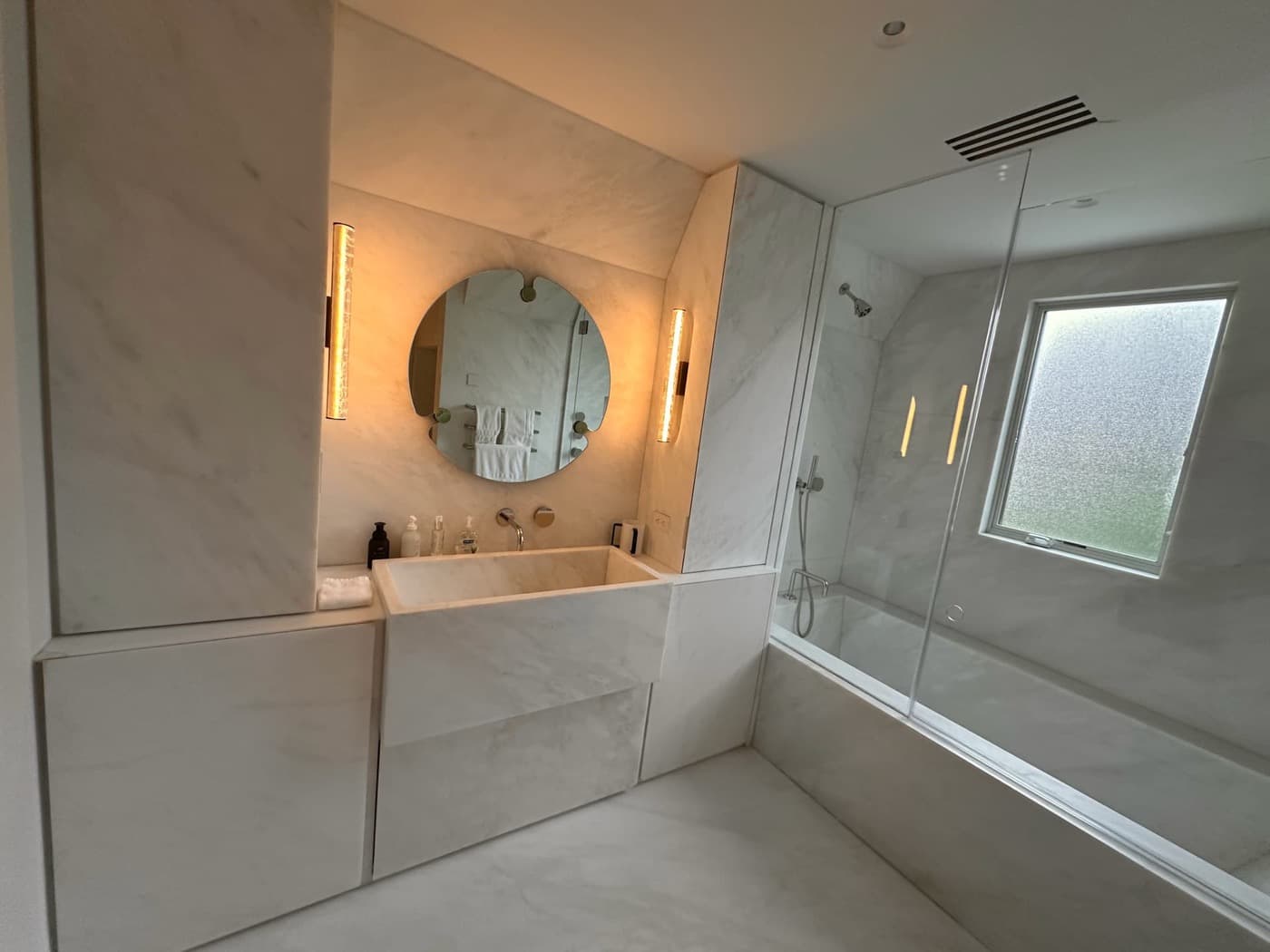 Bathroom clad in pale veined stone with integrated basin vanity, round mirror, and bathtub