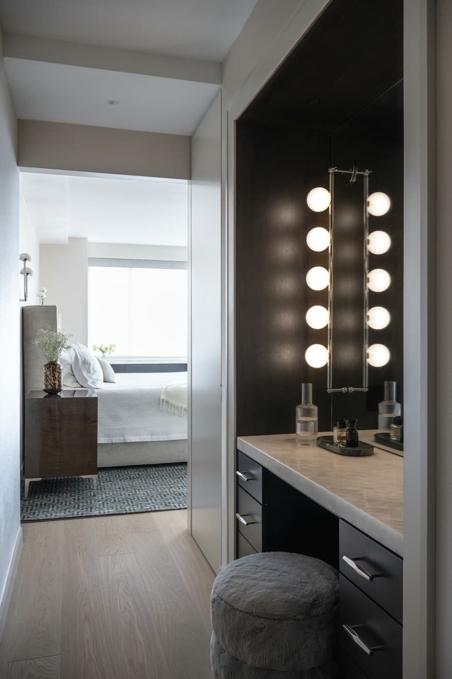 Built-in dark timber vanity alcove with illuminated mirror and pale stone countertop adjoining a bedroom