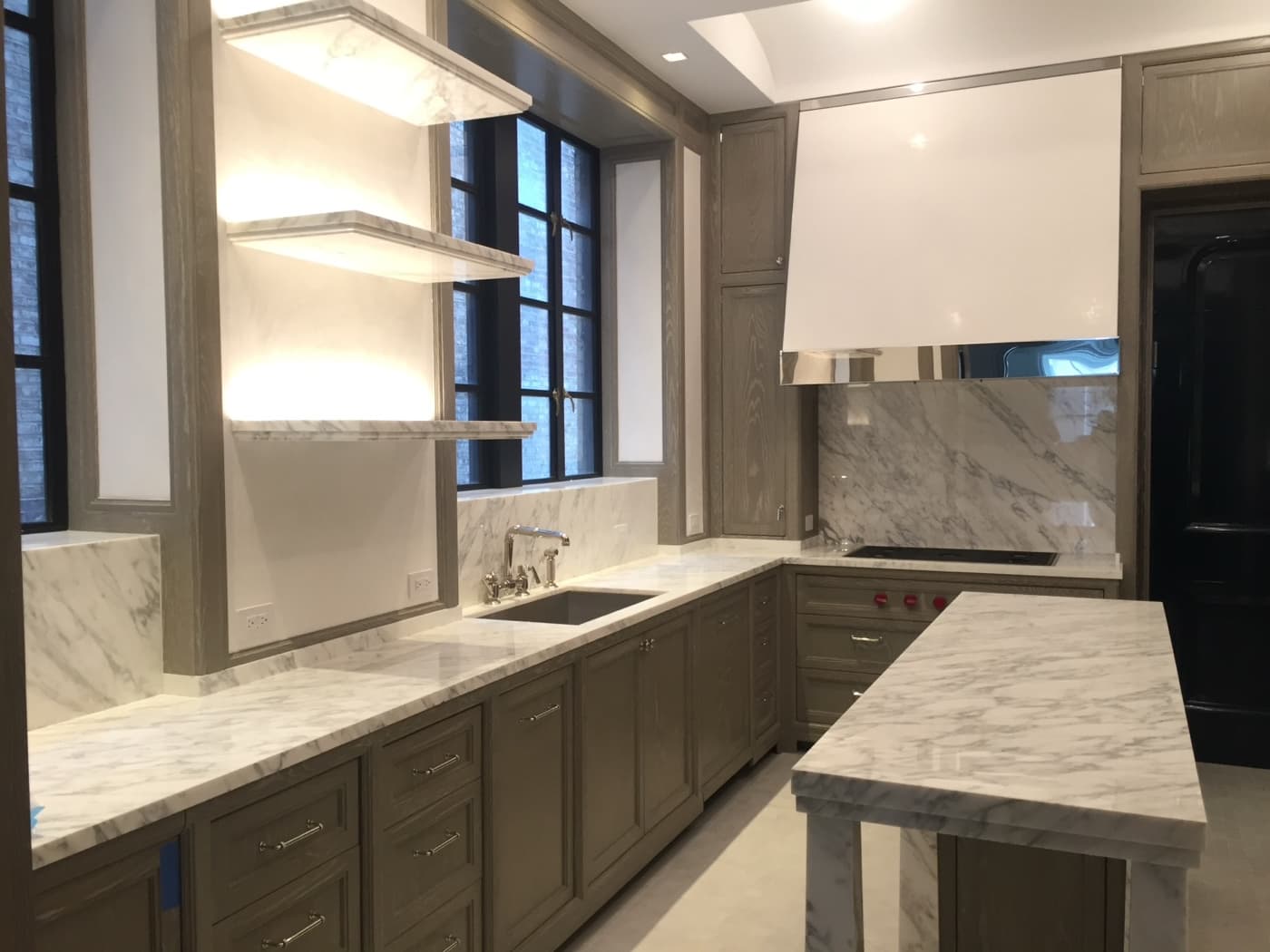 Kitchen featuring custom grey-toned millwork cabinetry, illuminated floating shelves, and pale veined stone surfaces
