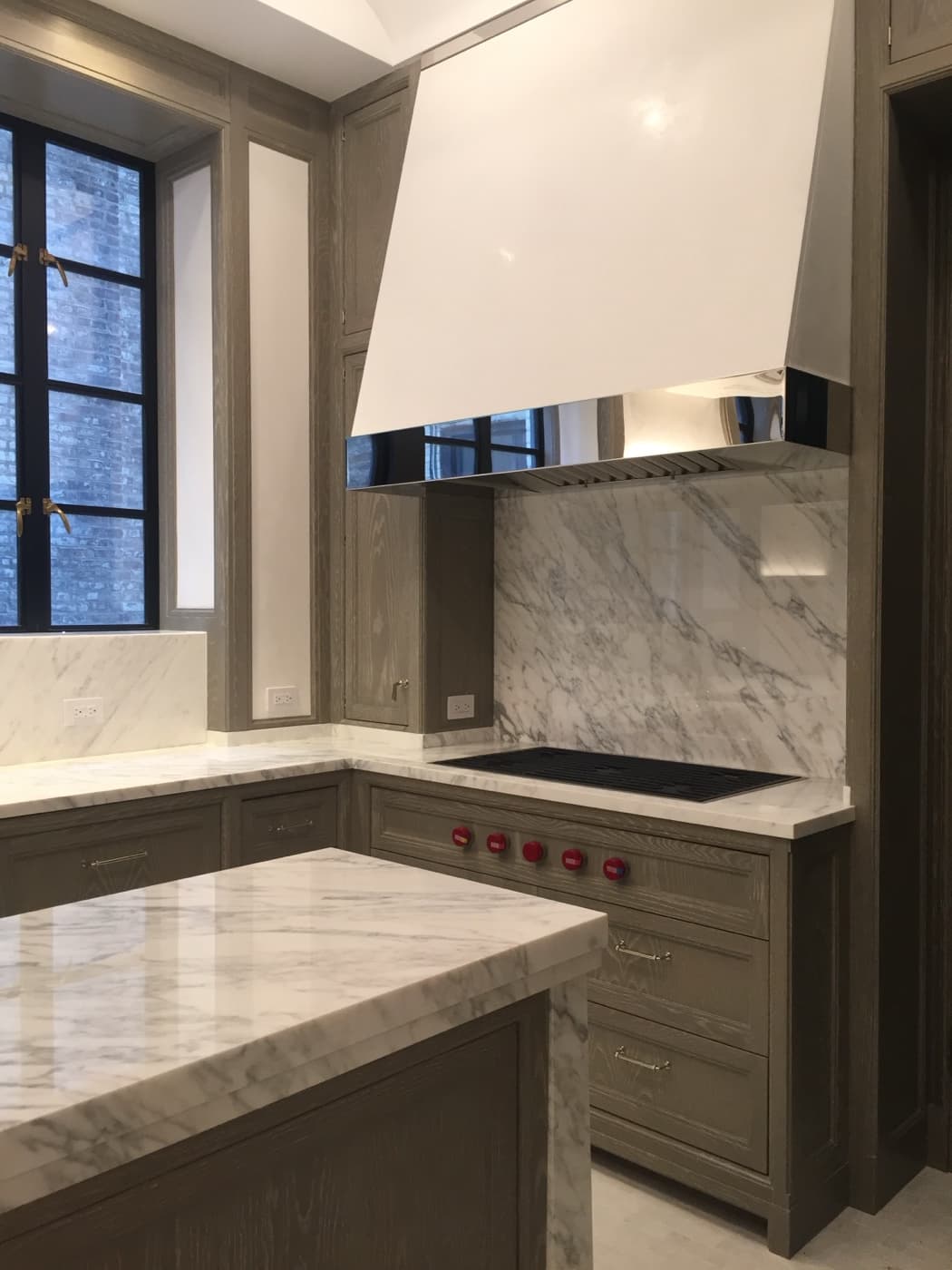 Kitchen with grey-toned wood cabinetry, pale veined stone surfaces, and a custom range hood
