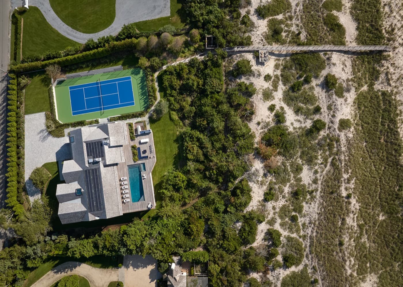 Coastal estate grounds with timber pool deck, blue tennis court, and beach boardwalk