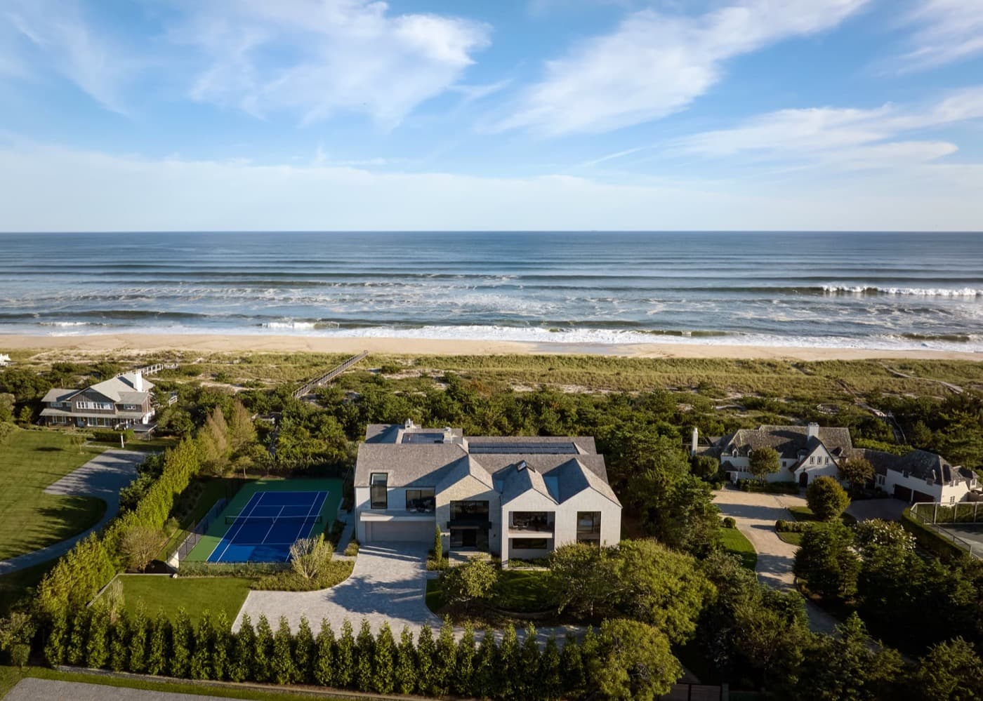 Coastal beachfront estate featuring modern gabled house, paved driveway, and private tennis court