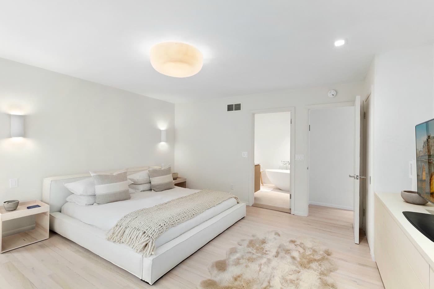 Minimalist bedroom featuring pale timber flooring, upholstered platform bed, and doorway into ensuite bathroom