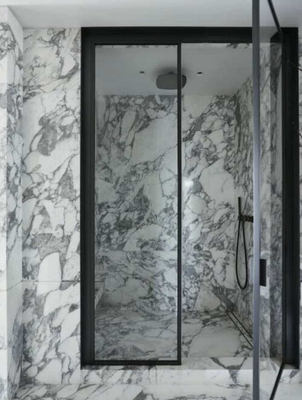 Walk-in shower clad entirely in dramatic veined pale stone with black metal-framed glass doors