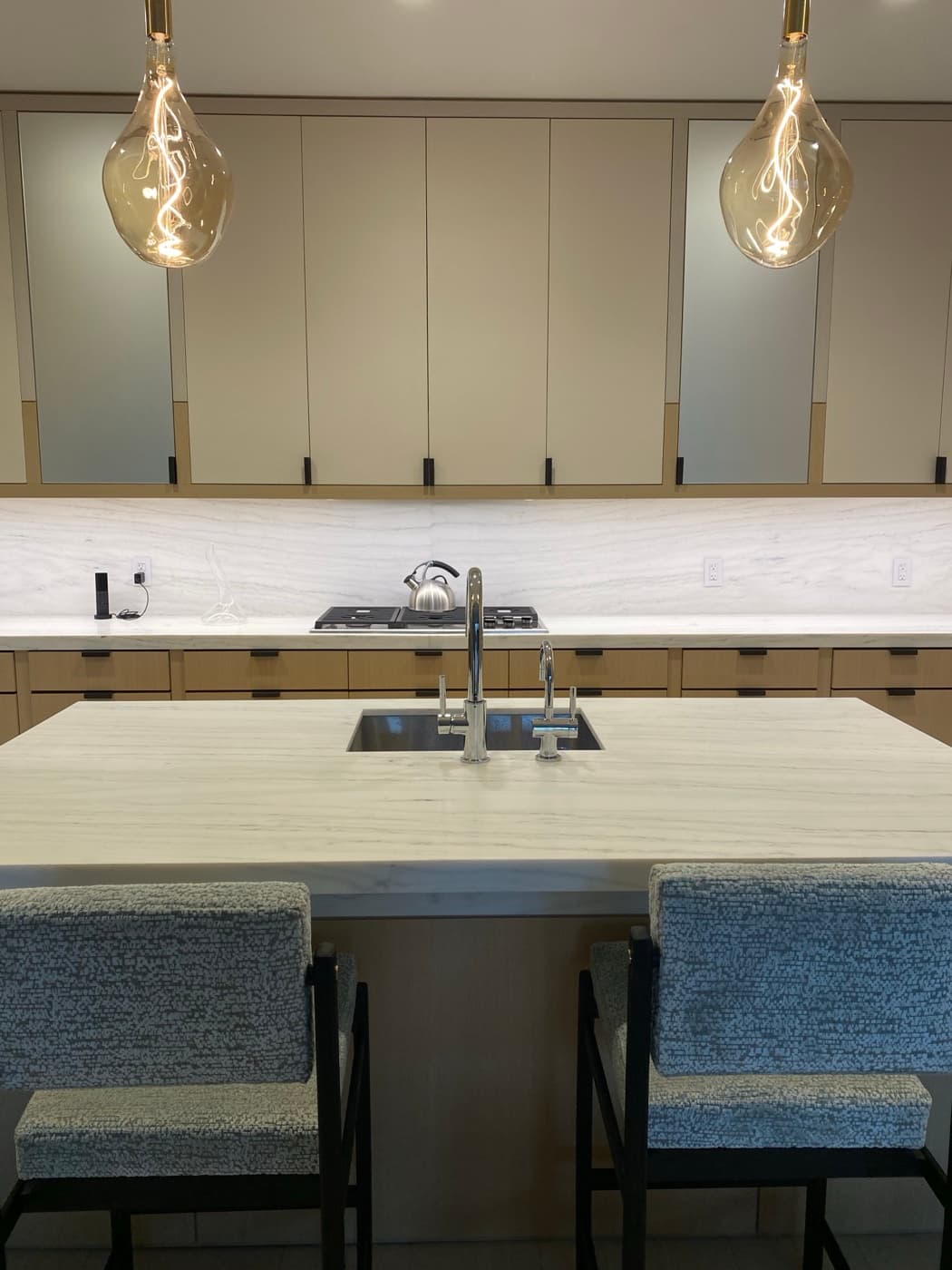 Kitchen featuring pale stone island, pendant lighting, and custom two-tone cabinetry with stone backsplash