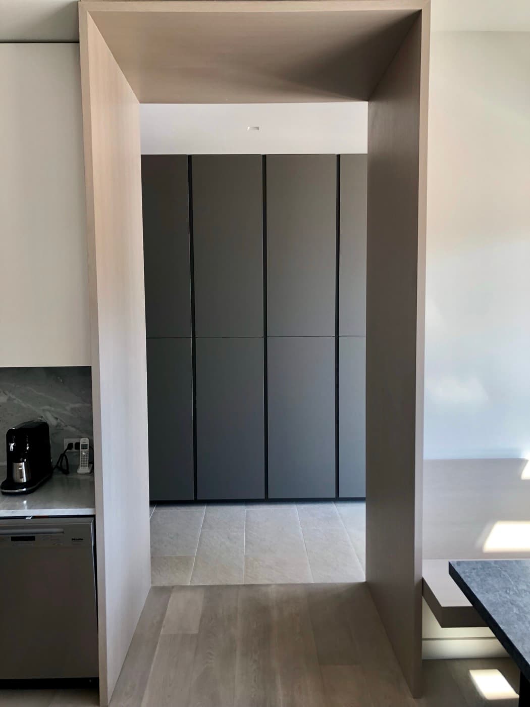 Pale timber portal connecting kitchen to hallway with full-height matte dark cabinetry