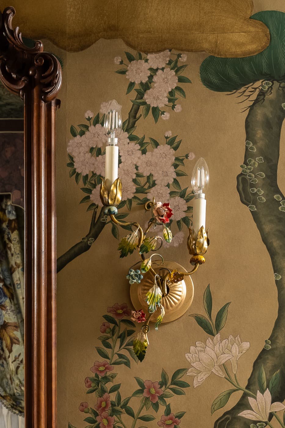 Bedroom detail with decorative floral sconce against botanical wallcovering beside an ornate carved timber frame