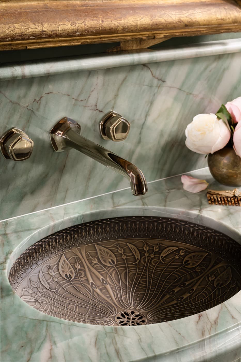 Bathroom vanity featuring green veined stone surfaces, wall-mounted faucet, and an ornate metal undermount basin