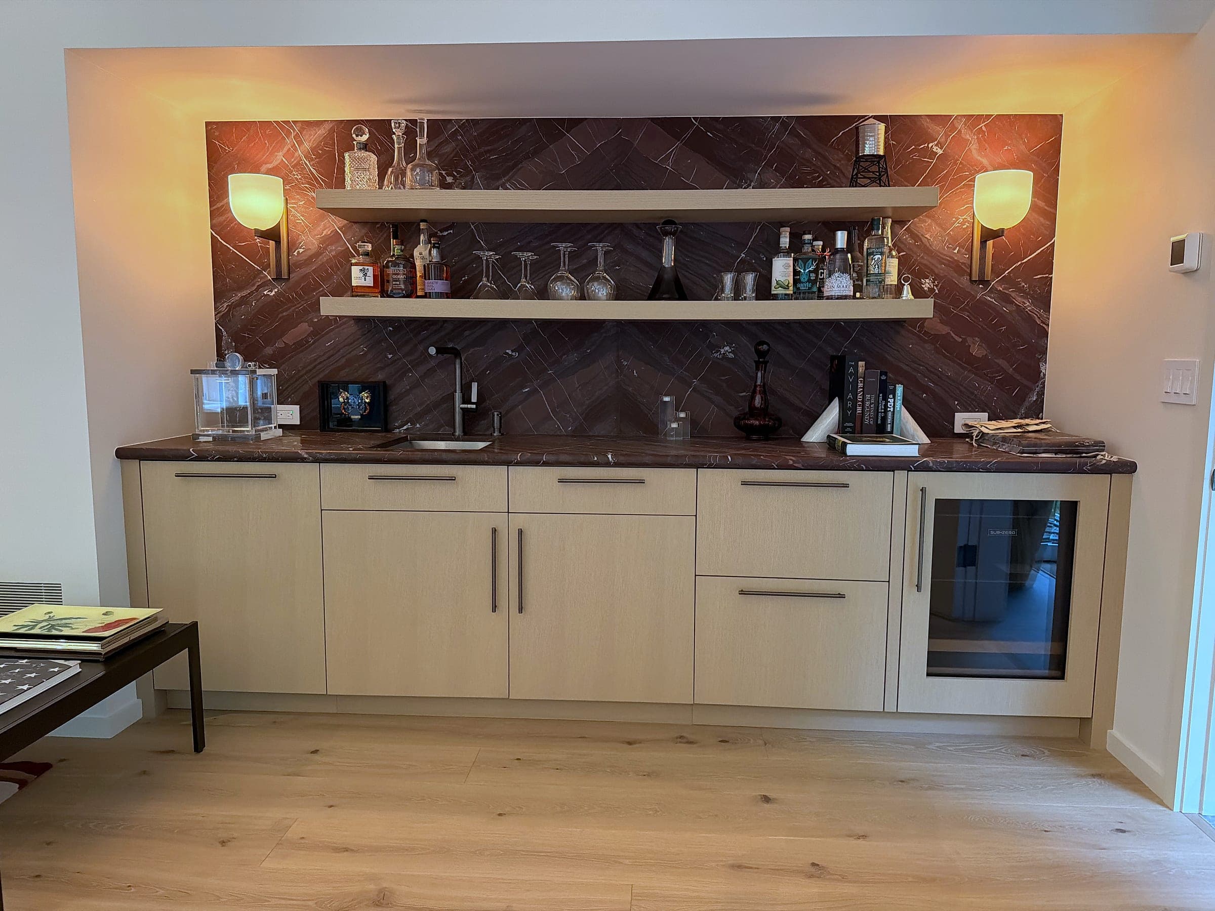 Wet bar run in rift-sawn white oak with a Rosso Levanto counter and herringbone backsplash, two floating shelves and wall sconces