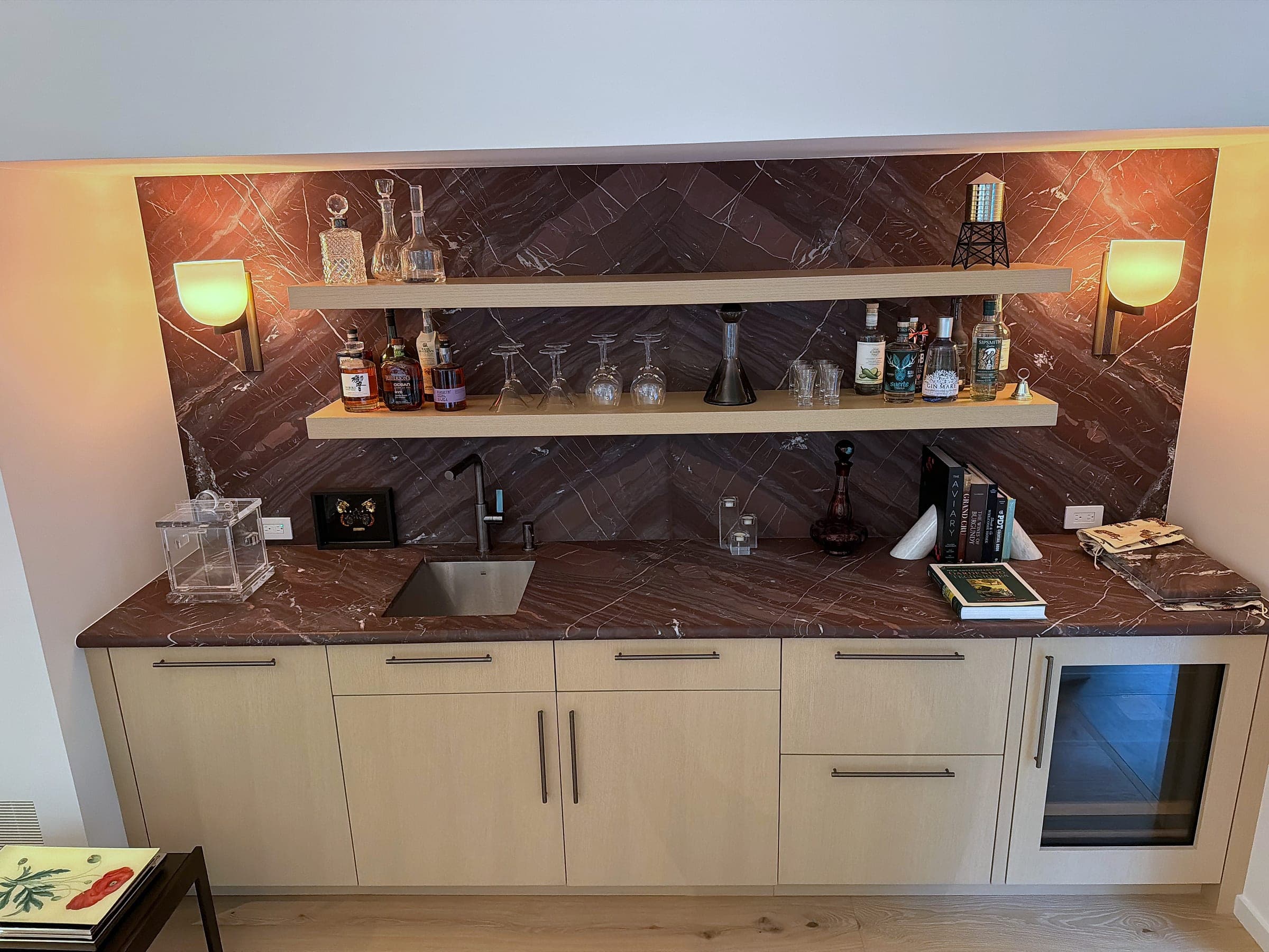 The wet bar seen at an angle, showing the undermount sink, drawer bank and glass-fronted undercounter wine store beneath the Rosso Levanto counter
