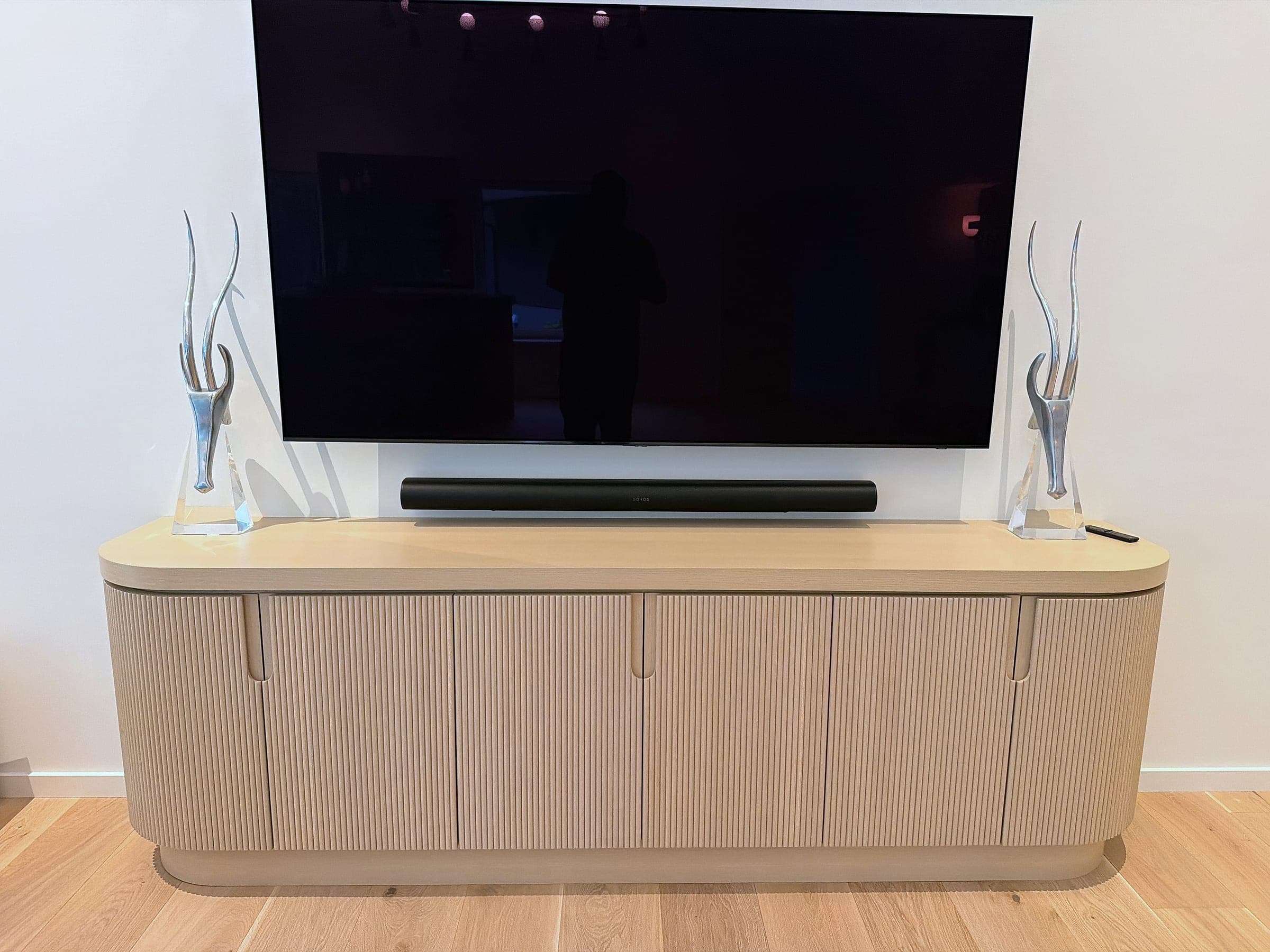 Fluted media credenza in pale oak with rounded ends, beneath a wall-mounted television